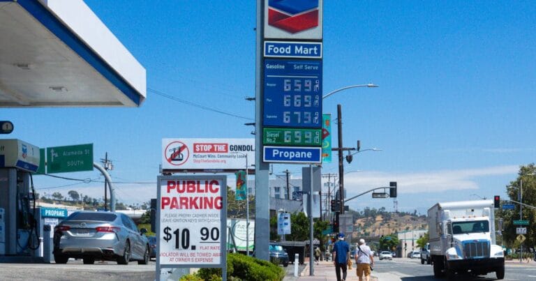 People walking near a Chevron gas station with prominent signage and parked vehicles on a sunny day.